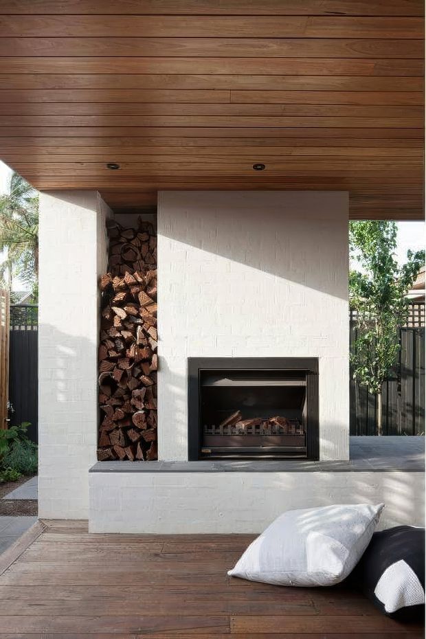 A compact outdoor entertaining area featuring a white-painted brick fireplace with an integrated firewood niche, set beneath a warm timber-lined ceiling. The space has timber decking, dark metal fencing, garden views, and loose outdoor cushions in the foreground. Strong sunlight creates a warm, relaxed atmosphere.