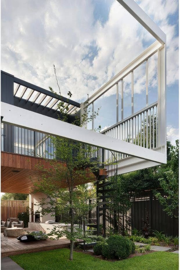 A contemporary multi-level residential courtyard viewed from outdoors, featuring dramatic white steel-framed glazing, projecting cantilevered volumes, a partially glazed roof structure, dark metal balustrades, and warm timber soffits and decking. A black spiral stair connects the levels, while landscaped planting, lawn, and outdoor seating occupy the courtyard below. The composition emphasises indoor-outdoor connection, transparency, and strong geometric architectural forms.