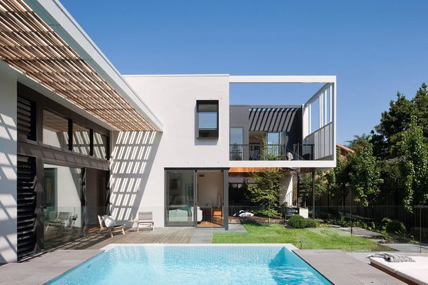 Exterior view of a contemporary multi-level residence organized around a backyard swimming pool. The white rendered facade has strong geometric forms, extensive glazing, dark-framed openings, and a projecting upper-level box with an open terrace. Timber slatted screens and pergola elements cast patterned shadows across the facade and paved pool terrace. Landscaped planting and lawn surround the pool in bright sunlight.