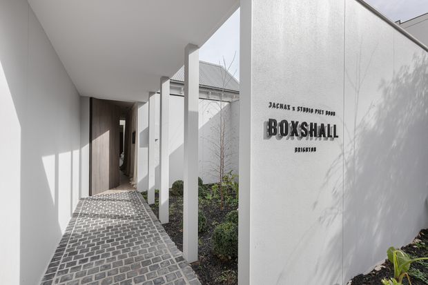A refined covered entrance walkway to Boxshall, featuring a white rendered facade, slim square columns, dark timber entry door, and a patterned grey brick-paved path bordered by landscaped planting. Black raised signage is mounted on the wall, with strong sunlight and crisp shadows creating a modern, polished arrival space.