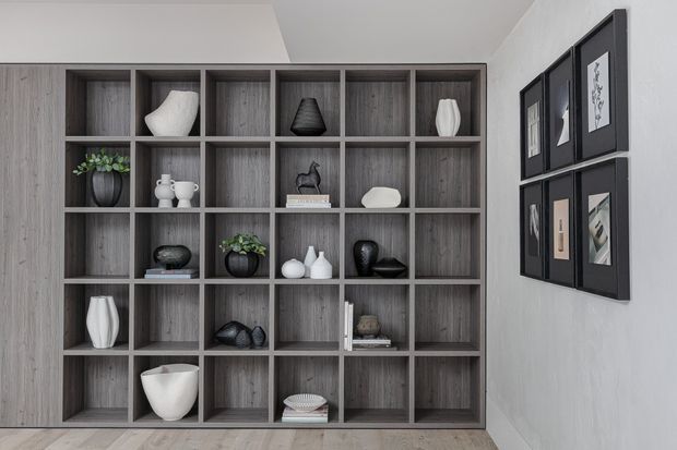 A contemporary interior wall featuring a large built-in open-grid shelving unit in grey wood grain, displaying monochromatic vases, sculptures, books, and potted plants. A coordinated arrangement of black-framed artwork hangs on the adjacent light wall. Light timber flooring and neutral finishes create a refined, gallery-like atmosphere.