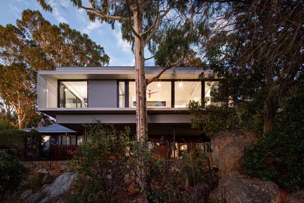 Exterior view of a contemporary two-storey residence partially screened by mature trees and landscaped rock outcrops. The upper level has a clean flat roofline, broad floor-to-ceiling windows, dark framing, and pale rendered wall surfaces. A covered lower-level outdoor living area and a small dark-roofed structure are visible beneath the elevated upper floor.