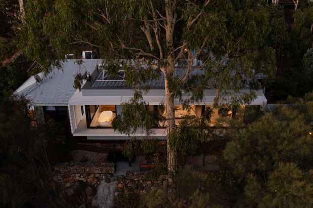 Aerial dusk view of a modern single-storey house nestled among dense native vegetation. The building has a low-pitched light metal roof, extensive glazing, a projecting balcony or terrace, and a rooftop skylight or glazed roof element. Warm interior lighting contrasts with the dark surrounding landscape and exposed rocky ground.