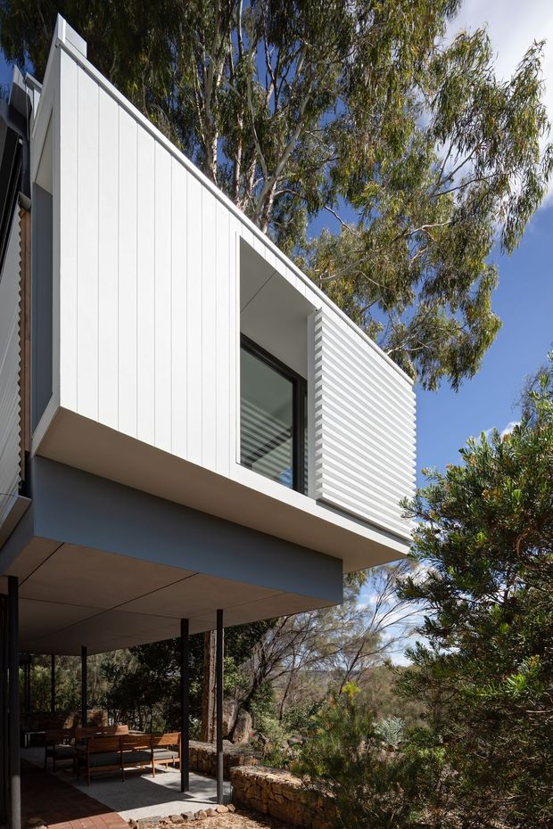Exterior architectural photograph of a contemporary elevated house set within a natural, wooded landscape. The building features crisp white vertical cladding, a projecting upper volume, a deep shaded covered outdoor living area, large glazing, and a horizontal louvred privacy screen. Stone retaining walls and native vegetation surround the structure under a clear blue sky.