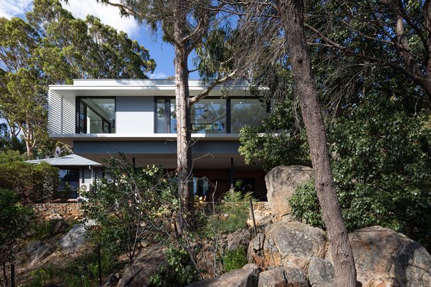 Exterior view of a contemporary two-storey house set among dense native vegetation and large exposed rocks. The building has a clean rectilinear form, a flat roof, extensive dark-framed glazing, white rendered walls, and a projecting upper level partially screened by trees.