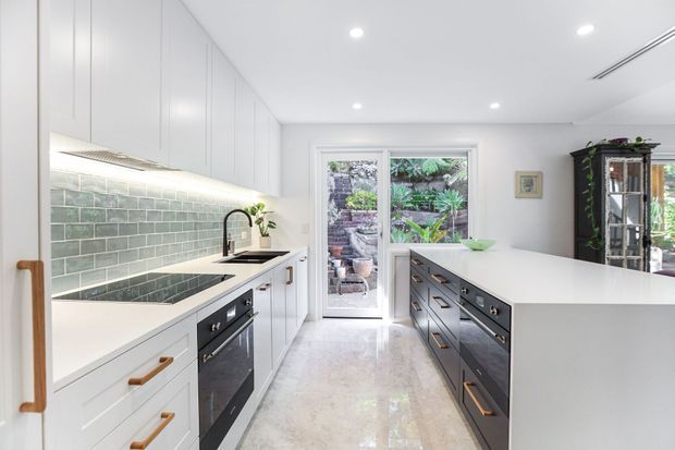 Bright contemporary galley-style kitchen with white full-height cabinetry, dark lower cabinetry, pale stone benchtops, a muted green tiled splashback, integrated appliances, and a central island. A glazed door and window open to a lush garden, while a tall glass-fronted cabinet adds storage at the rear. Recessed ceiling lighting and under-cabinet illumination create a clean, airy atmosphere.