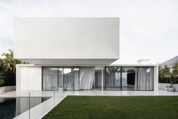 A minimalist contemporary two-storey residence with a stark white cubic upper volume projecting over a fully glazed ground floor. The facade features extensive floor-to-ceiling glazing, sheer curtains, white walls, and a crisp linear composition. A transparent glass pool fence borders a reflecting or swimming pool beside a manicured lawn, with tropical planting and neighbouring roof forms visible in the background.
