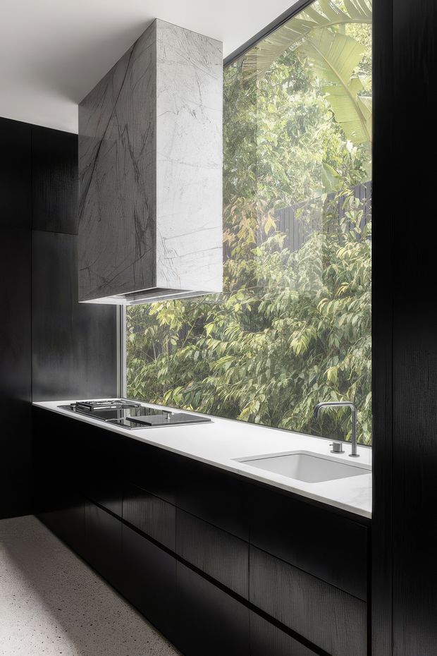 A minimalist contemporary kitchen with a dramatic black fitted cabinet wall and island-style run of cabinetry, a bright white stone-look benchtop, integrated cooktop, undermount sink and slim mixer tap. A large floor-to-ceiling window brings in abundant natural light and overlooks dense greenery. The kitchen has a clean, high-contrast palette and refined, pared-back detailing.