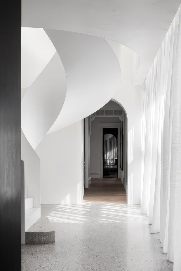 A bright, minimalist interior hallway with a sculptural curved white ceiling and wall treatment, arched doorways leading through a series of rooms, sheer floor-to-ceiling curtains along the right side, and a dark vertical timber or wood-toned wall panel at left. The foreground floor appears light terrazzo or polished concrete, while the deeper corridor has dark timber flooring. Strong sunlight creates linear shadows across the floor.