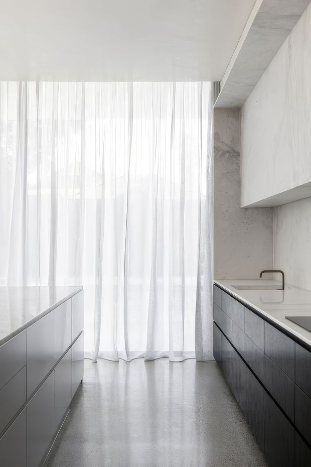 A bright, minimalist contemporary kitchen with a central island, dark linear cabinetry, pale benchtops, and a large wall of sheer white curtains providing soft daylight. The space features a polished grey floor, concrete-look wall and ceiling surfaces, integrated appliances, and a simple bronze-toned tap.