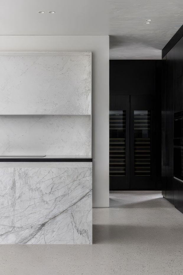 A refined monochromatic kitchen interior featuring a large white veined stone island or benchtop in the foreground, dark integrated cabinetry and wine storage at the rear, black vertical wall joinery, a smooth light ceiling with recessed downlights, and a pale terrazzo-like floor. The composition emphasizes clean lines, contrasting light stone and near-black cabinetry, and a minimalist high-end fit-out.