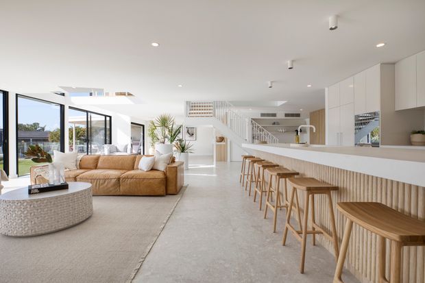 Bright open-plan contemporary interior combining a living room and kitchen. A large white island with integrated seating and vertical timber detailing dominates the right side, while a tan sectional sofa, round coffee table, expansive glazing, polished light concrete flooring, and a staircase create an airy, spacious setting.
