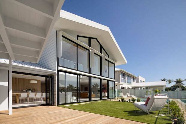 Bright exterior view of a contemporary multi-level home with a strongly glazed black-framed facade, white horizontal cladding, projecting roof forms and extensive outdoor living areas. A timber deck leads to a manicured lawn, outdoor lounge seating and a swimming pool, while the open dining and kitchen area is visible through the glazing.