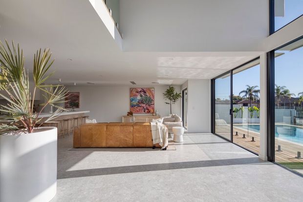 Bright, spacious open-plan living area with a large tan sectional sofa, white furnishings, a kitchen and island in the background, and full-height glazed sliding doors opening to a pool terrace. A double-height void and clean white surfaces create an airy contemporary interior, with tropical planting and pool views outside.