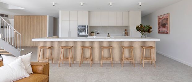Bright open-plan kitchen and living area with a long white island bench, six timber bar stools, white flat-panel cabinetry, integrated appliances, pale stone-look surfaces, and a vertically slatted timber island front. A staircase and partial tan leather seating are visible at left, with neutral carpeted flooring, recessed ceiling lights, and restrained artwork and greenery.