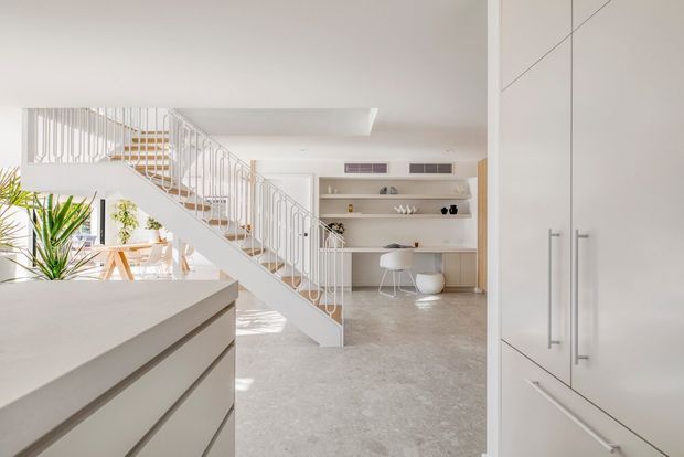 Bright, open-plan contemporary interior viewed from a kitchen, with pale built-in cabinetry in the foreground, a sculptural white staircase with slender balusters, and a light-toned living or dining area beyond. Built-in shelving and a compact desk workspace sit beneath concealed air-conditioning vents, while indoor plants and warm timber details add subtle natural accents. The space has a clean, airy, minimalist appearance with a polished light stone or concrete-look floor.