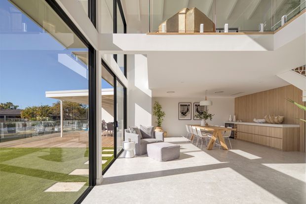 Bright, double-height contemporary open-plan living space with a dining area and linear kitchen. Floor-to-ceiling black-framed glazing opens to a landscaped outdoor area, while a mezzanine level overlooks the room. The interior features pale finishes, a timber-look vertical feature wall and cabinetry, a large neutral sectional sofa, and strong sunlight creating geometric shadows across the polished floor.