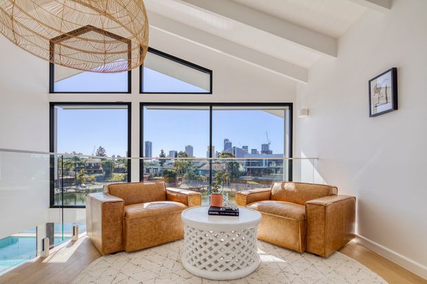 Bright contemporary living room with two tan leather armchairs, a white sculptural round coffee table, and a large pale rug. Floor-to-ceiling black-framed glazing overlooks a waterfront-like urban neighbourhood and skyline, while a glass balustrade borders an open stair or lower-level void. Exposed white ceiling beams and a large woven pendant add architectural and natural-textured detail.