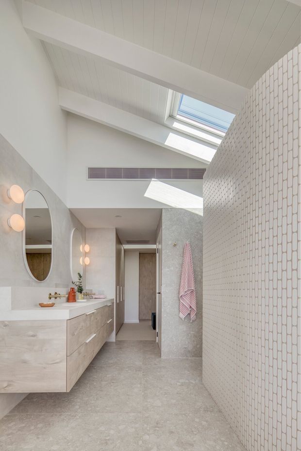 Bright contemporary bathroom with a floating pale timber vanity, white benchtop, oval mirror, wall-mounted globe lights, concrete-look walls and floor, and a full-height white vertically patterned tile feature wall. A skylight and high pitched white panelled ceiling bring in abundant natural light. The room includes a shower area screened by the tiled wall, open shelving, and a pink striped towel.