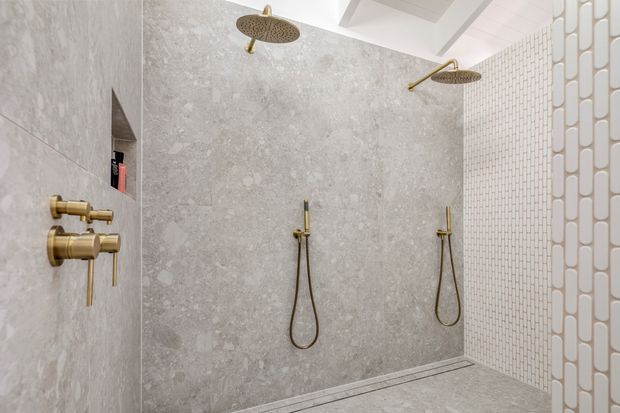 Luxurious walk-in shower with dual rainfall showerheads and separate handheld fittings, finished in large-format pale grey stone-look wall and floor tiles. A white small-format tiled feature wall and decorative glazed screen frame the space, while brushed brass fixtures add warmth. The ceiling includes recessed beams or a coffered detail.