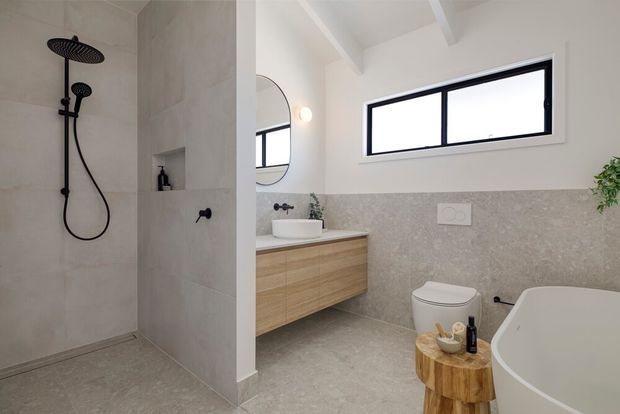 Bright contemporary bathroom with a walk-in tiled shower, black rain-shower fittings, floating timber vanity with a white vessel basin, wall-mounted mirror, freestanding bathtub, toilet, and high horizontal window. Pale stone-look tiles and white walls create a clean, neutral interior with black accents and natural timber detailing.