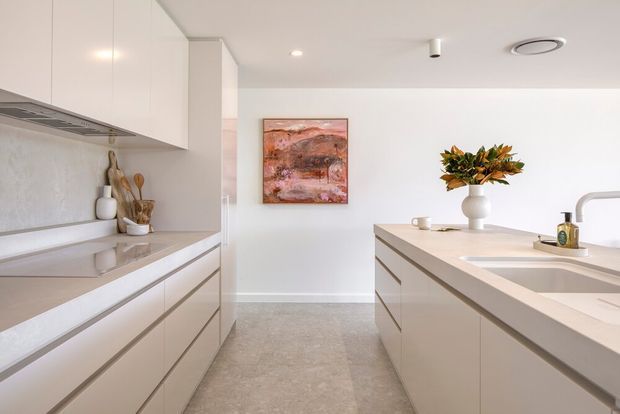 Bright contemporary kitchen with a central island, pale flat-panel cabinetry, integrated appliances, light stone benchtops, and a textured neutral splashback. The space opens toward a dining or living area, with minimalist styling, recessed lighting, a linear pendant-free ceiling, and a warm abstract artwork and vase arrangement providing subtle colour.