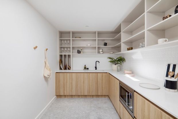 Bright, minimalist kitchen with an L-shaped run of light timber-grain cabinetry, white benchtops, white tiled splashback, open shelving, integrated appliances, and a black tap and sink. The compact room has pale walls and a light grey floor, creating a clean, airy contemporary interior.