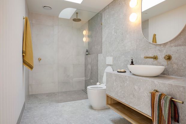 Contemporary bathroom with a walk-in glass shower, wall-mounted rain showerhead, wall-hung toilet, vessel basin on a floating vanity, large round mirror, integrated lighting, and continuous pale grey stone-look wall and floor surfaces. Warm brass tapware and timber-toned open vanity shelving add contrast, while striped towels provide a small decorative accent.
