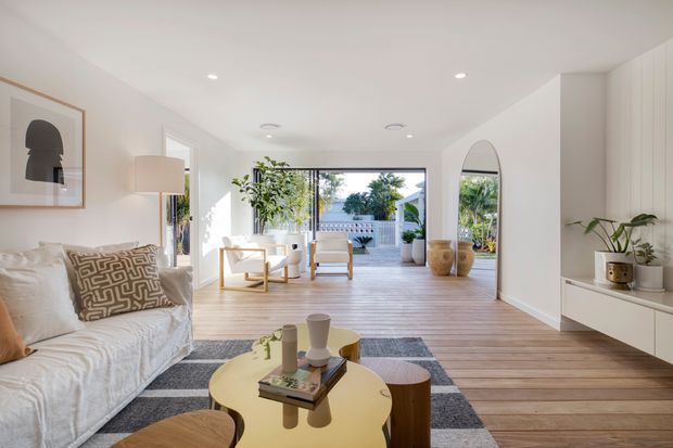 Bright, open-plan living room with white walls, pale timber flooring, a linen sofa, round coffee table, neutral rug, sculptural decor, built-in white cabinetry, indoor plants, and large glazed openings leading to an outdoor terrace or courtyard. The room has a clean contemporary coastal feel with abundant natural light.