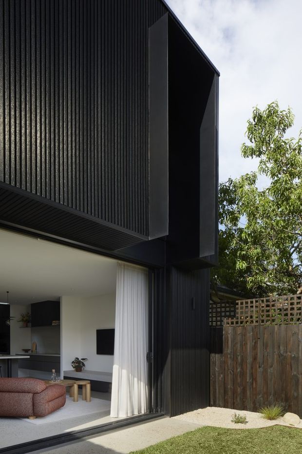 Contemporary residential exterior showing a dramatic black vertically ribbed upper facade and projecting box form over a highly glazed living area. The open corner reveals a minimalist living room with a rust-coloured sectional sofa, television, dark built-in cabinetry, pale flooring, and sheer curtains. A small landscaped garden, timber boundary fence, and lattice screen sit beside the house.