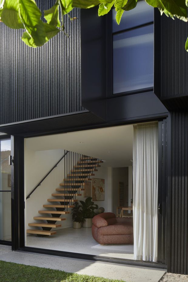 A contemporary multi-level home viewed from the garden through a large glazed opening. The dark vertically ribbed exterior cladding frames a recessed living area with a floating timber stair, slender black balustrade, pale terrazzo-like flooring, white curtains, indoor planting, and a rounded rust-coloured lounge chair. An upper projecting volume contains a frosted or translucent window, while leafy vegetation partially frames the view.