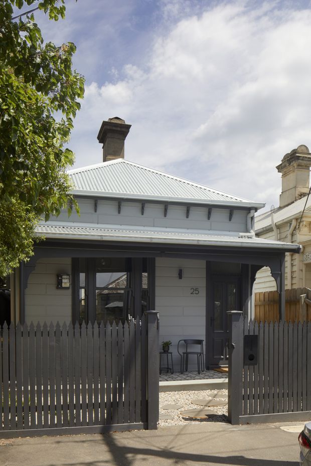 Street-facing view of a compact heritage-style weatherboard dwelling with a pale grey corrugated metal roof, prominent chimneys, decorative eaves brackets, and a covered front porch. A dark grey picket fence and gate enclose a small gravel and stepping-stone entry courtyard. The facade combines traditional detailing with a restrained contemporary grey-and-white colour scheme.