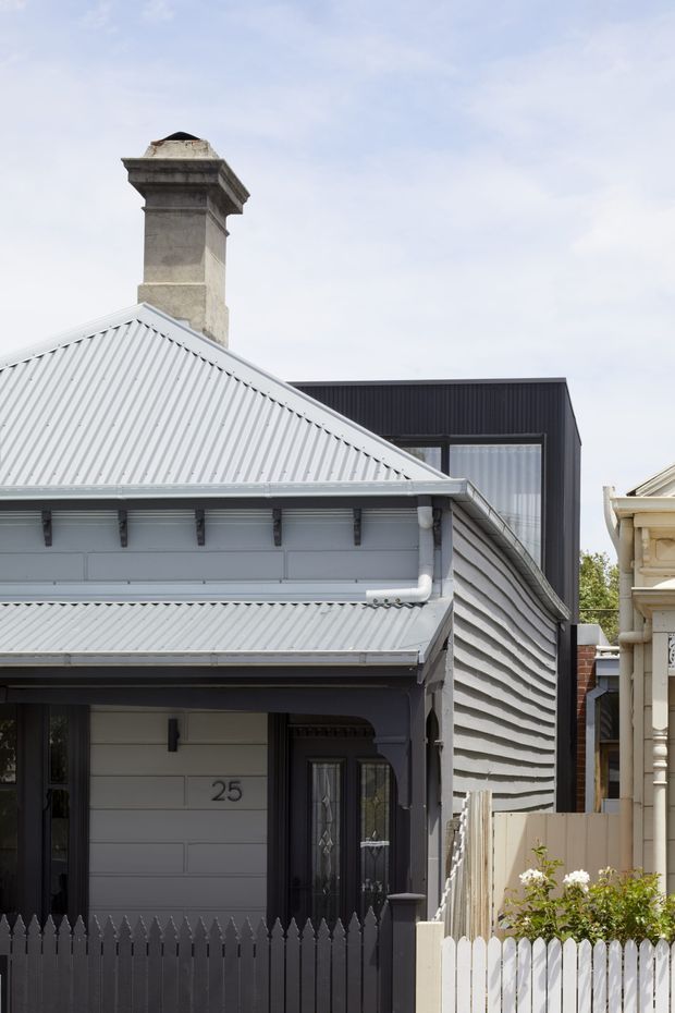 Street-facing view of a compact heritage-style weatherboard house with a steep corrugated metal gable roof, ornate masonry chimney, timber eaves detailing, front porch entry, and a dark grey picket fence. A contrasting contemporary dark-clad extension with large glazing rises behind the original dwelling, while part of a neighbouring traditional facade is visible at right.