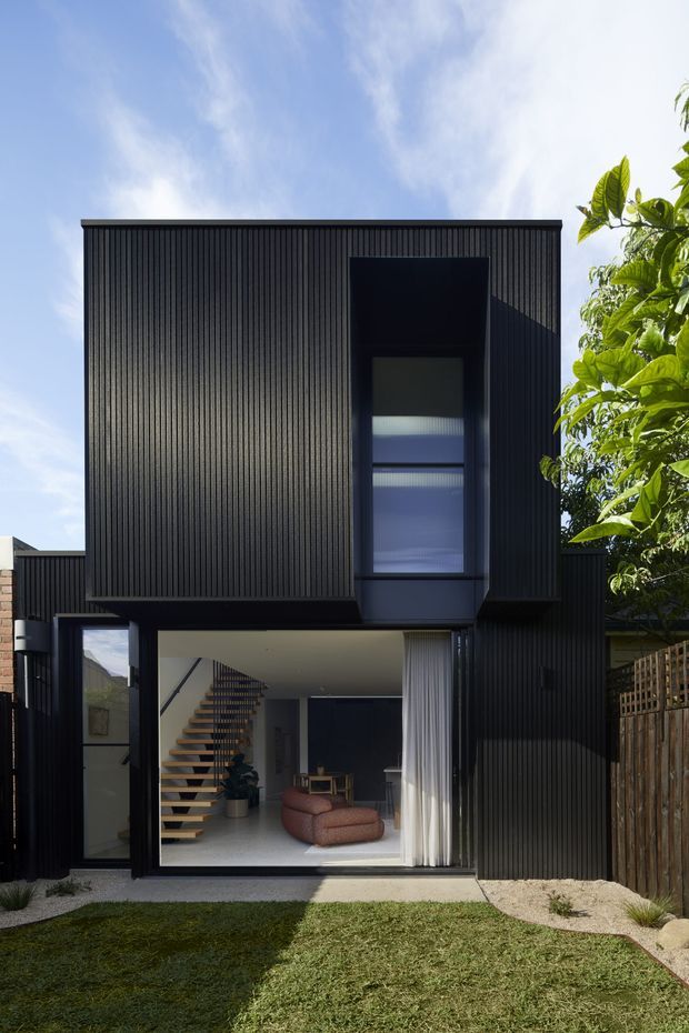Front exterior view of a contemporary two-storey residence with a bold black vertically ribbed facade. A tall recessed window projects slightly from the upper level, while the ground floor opens through large glazed doors to a minimalist living area with a floating timber staircase, pendant lighting, and a low upholstered sofa. A small lawn, gravel edging, timber boundary fencing, and mature planting frame the house.