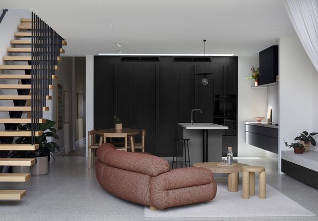 A contemporary open-plan living area with a sculptural curved sofa facing a compact round coffee table, a minimalist kitchen with a central island, and a dining table beyond. A floating timber staircase with a black vertical balustrade occupies the left side, while full-height dark cabinetry forms a dramatic feature wall. Light polished concrete-style flooring, white walls and ceilings, and soft neutral furnishings create a refined, modern interior.