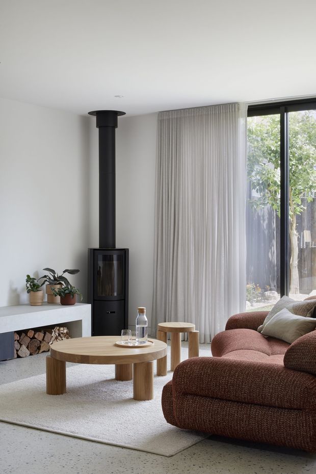 A bright, contemporary living room with a low white ceiling, white walls, full-height sheer curtains, and black-framed glazing. A sculptural black wood-burning stove sits beside a white fireplace hearth with stacked firewood beneath. The room is furnished with a large textured rust-brown sectional sofa, a round light timber coffee table, matching side table, neutral cushions, indoor plants, and a pale textured rug over a light speckled floor.