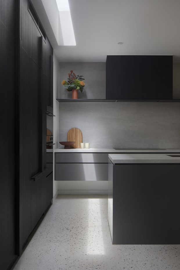 A refined, compact contemporary kitchen with tall dark timber cabinetry on the left, a dark island in the foreground, streamlined grey cabinetry, and a pale grey stone or concrete-look backsplash. A long skylight brings soft natural light into the space. The floor appears to be light terrazzo, while a small arrangement of flowers and timber serving boards adds warmth to the otherwise monochromatic scheme.