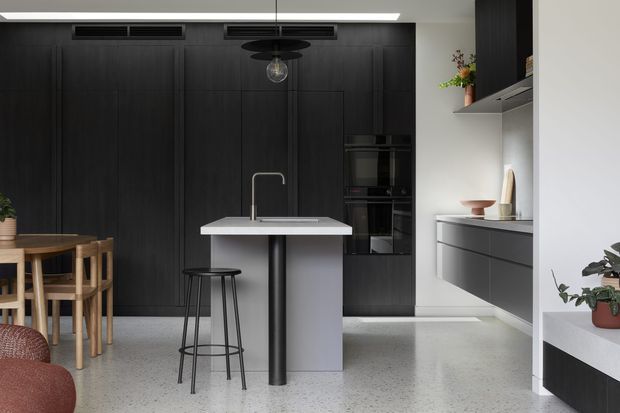 A sophisticated contemporary kitchen with a compact central island, integrated sink and black tap, dark full-height cabinetry, built-in ovens, and a light grey terrazzo-style floor. A timber dining table and chairs sit to the left, while open shelving, dark cabinetry, and restrained decorative objects line the right wall. The room has a high-contrast black, white, and grey palette with warm timber accents and a pendant light above the island.