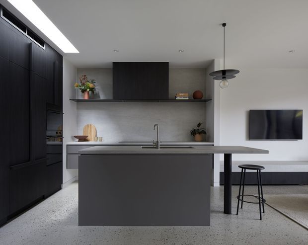 A refined open-plan kitchen with a large dark grey island, integrated sink and slim stone benchtop. Full-height matte black cabinetry lines the left wall, while a grey textured splashback, open timber shelf, black pendant light, and a TV area with bar stool are visible to the right. The terrazzo-style floor and restrained monochromatic palette create a contemporary, minimalist interior.