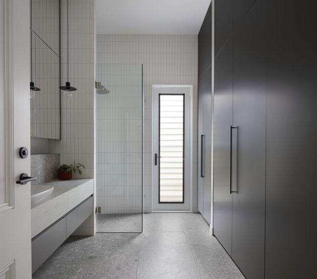 Contemporary bathroom with a walk-in glass shower, wall-mounted double vanity, large mirrored cabinet, pale vertically tiled walls, grey stone-look floor tiles, and a narrow frosted louvre-style window. Dark charcoal built-in cabinetry lines the right wall, creating a clean high-contrast scheme.
