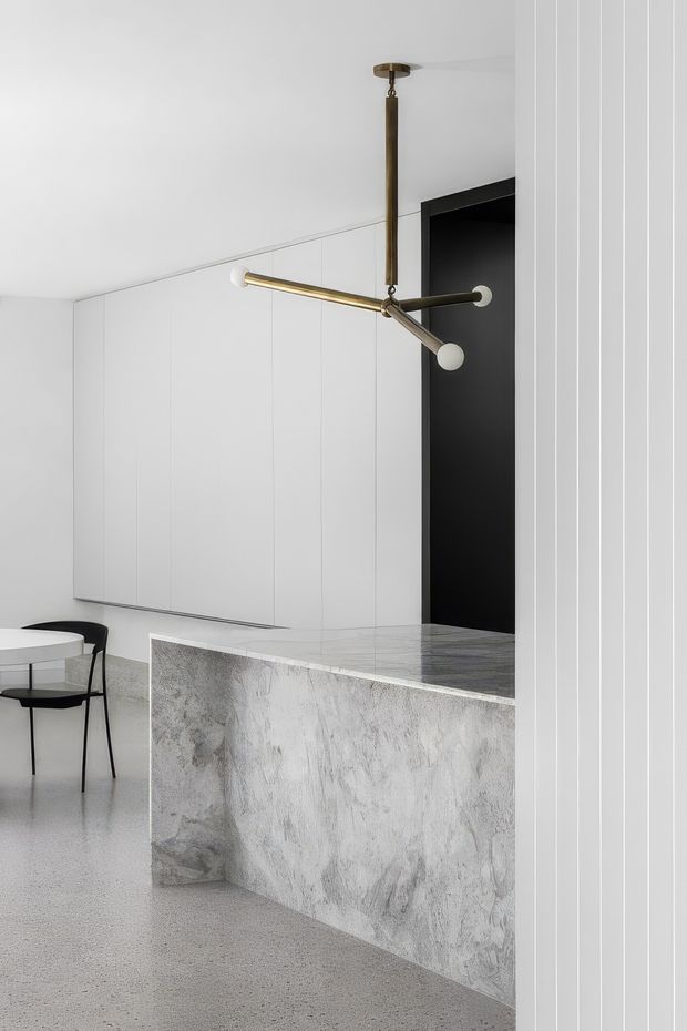 Minimal, highly refined kitchen interior featuring a monolithic pale grey stone island with waterfall-style side panel, integrated white vertical-panel cabinetry, a black recessed doorway or niche, and a sculptural aged-brass multi-arm pendant light. A small round dining table and black chairs sit at the edge of the space. The room has a restrained monochrome palette with subtle textural contrast and a polished contemporary fit-out.