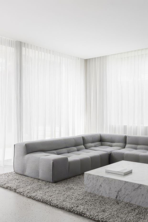 A bright, minimalist living room with a large low-profile grey modular sectional sofa arranged around a substantial white veined stone coffee table. Floor-to-ceiling sheer white curtains wrap around the corner glazing, while a textured grey rug and pale floor create a soft monochromatic palette.