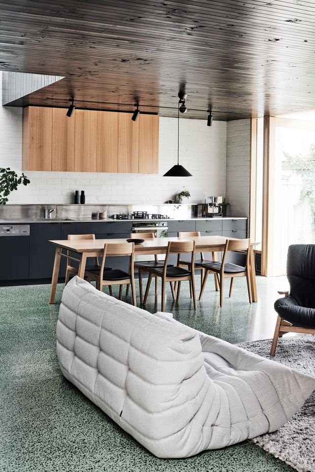 Open-plan interior combining a contemporary kitchen, dining area and living space. The kitchen has dark charcoal cabinetry, a stainless-steel benchtop, white painted brick walls, and a warm timber overhead cabinet. A timber dining table with mid-century style chairs sits between the kitchen and living area. The room features a heavily timber-lined ceiling, terrazzo-like green flooring, large glazing, a sculptural white lounge chair, and a black pendant light.