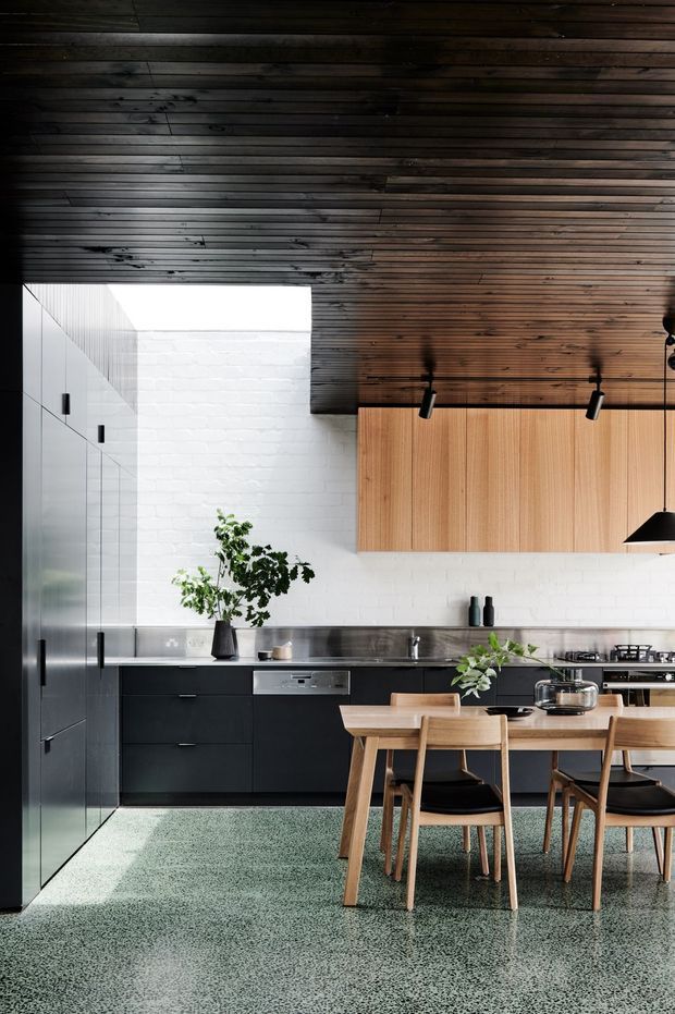 A stylish open-plan kitchen and dining area with dark lower cabinetry, full-height integrated storage, a stainless-steel benchtop, white painted brick walls, and warm timber overhead cabinetry. The ceiling is clad in dark timber slats, while a large terrazzo floor extends through the room. A timber dining table with matching chairs sits in the foreground, complemented by black pendant and track lighting.
