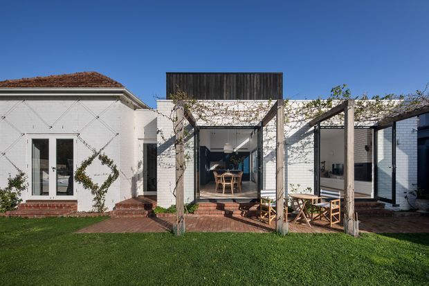 Front-facing exterior view of a low-rise residential home with a white-painted brick facade, red tiled roof, and a series of glazed openings leading to a brick-paved courtyard or terrace. Timber pergola structures with climbing vines frame the outdoor dining area, while dark vertical timber cladding forms a raised feature volume above the central section. A neat lawn occupies the foreground under clear daylight.