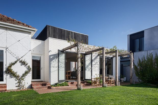 Exterior view of a contemporary residential extension or courtyard composition, combining an older white-painted masonry building with a tiled roof and newer white brick volumes. A timber pergola with climbing vines shelters a small outdoor dining area, opening onto a manicured lawn. Dark vertical timber cladding and vertical screening add contrast to the modern additions.