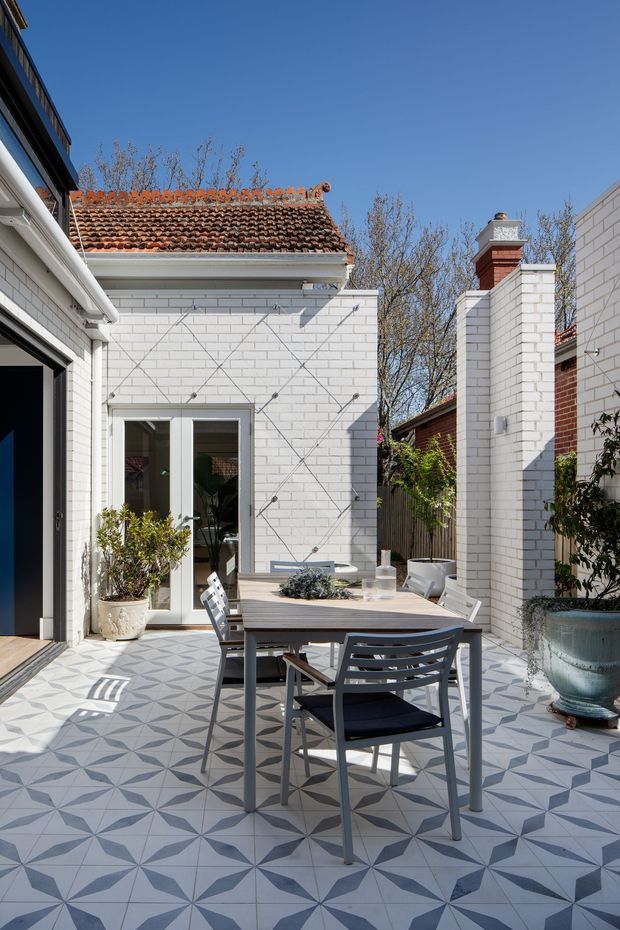 A bright, enclosed urban courtyard or terrace with patterned geometric floor tiles, a modern outdoor dining table and chairs, large planters, and white-painted brick walls. The space combines a contemporary addition with a traditional terracotta-tiled roof and chimney visible above the courtyard walls.