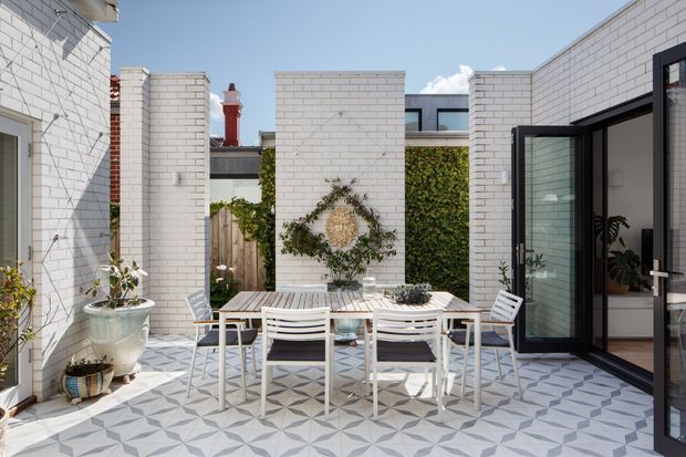 Bright courtyard terrace with white brick walls, patterned geometric tiled paving, a white outdoor dining table and chairs, potted plants, and black-framed glazed doors opening into the home. A vine-covered garden wall and decorative wreath add greenery and texture.