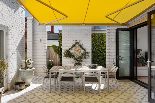 A bright, enclosed courtyard-style outdoor dining terrace with white brick walls, patterned tiled flooring, a white dining table and chairs, potted plants, and a large yellow retractable shade canopy. Dark-framed glazed doors open into the adjoining interior, while climbing greenery and a timber privacy fence add texture.
