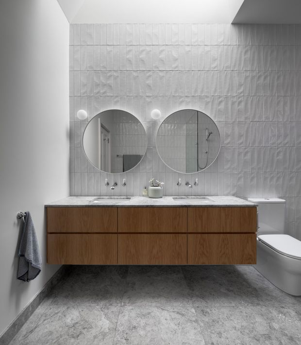 A contemporary bathroom featuring a wall-to-wall floating timber vanity with two integrated white basins, round mirrors, wall-mounted lights, and a white toilet. The walls are clad in textured white rectangular tiles, while the floor has a grey stone-like finish. A shower is visible in the mirror reflection.